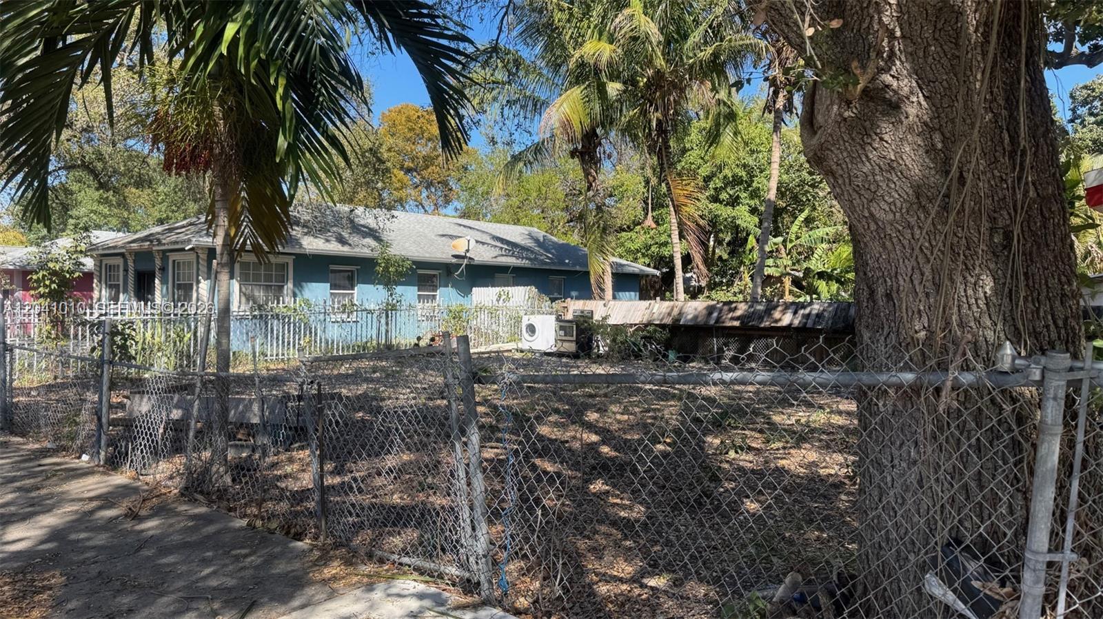A blue house sits behind a chain-link fence with a large tree in the foreground — 7613 NE 3rd Ave, Miami FL 33138, MIAMI, FL