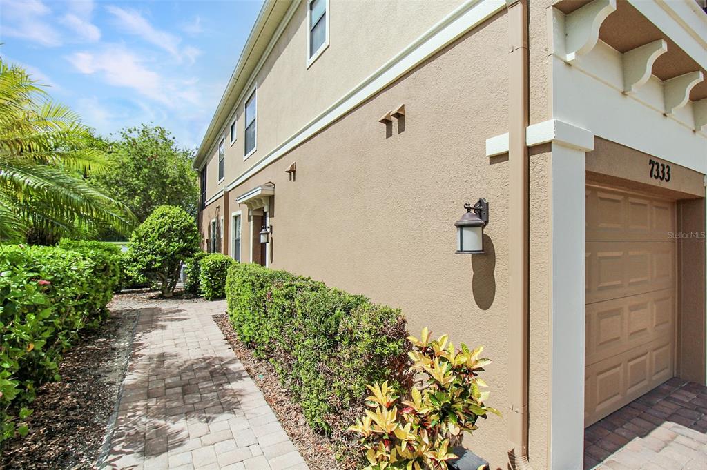 A tan exterior wall with a garage door, a brick walkway, and trimmed green hedges — 7333 SKYBIRD ROAD, BRADENTON, FL