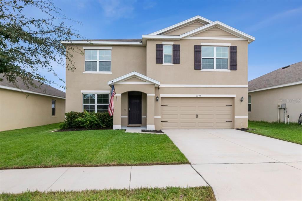 A two-story suburban house with a tan stucco exterior, a two-car garage, and a lawn — 6803 EZRA LOFT PLACE, ZEPHYRHILLS, FL