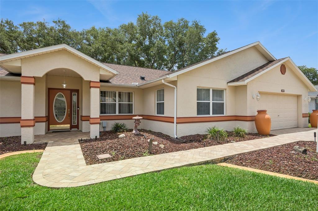 A single-story house with a beige stucco exterior, brown roof, and a paved walkway — 25904 NEWCOMBE CIRCLE, LEESBURG, FL