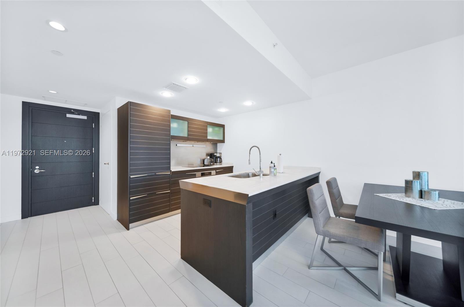 A modern kitchen features dark wood cabinetry, a white countertop island, and gray dining chairs — 200 Biscayne Boulevard Way # 3811, Miami FL 33131, MIAMI, FL