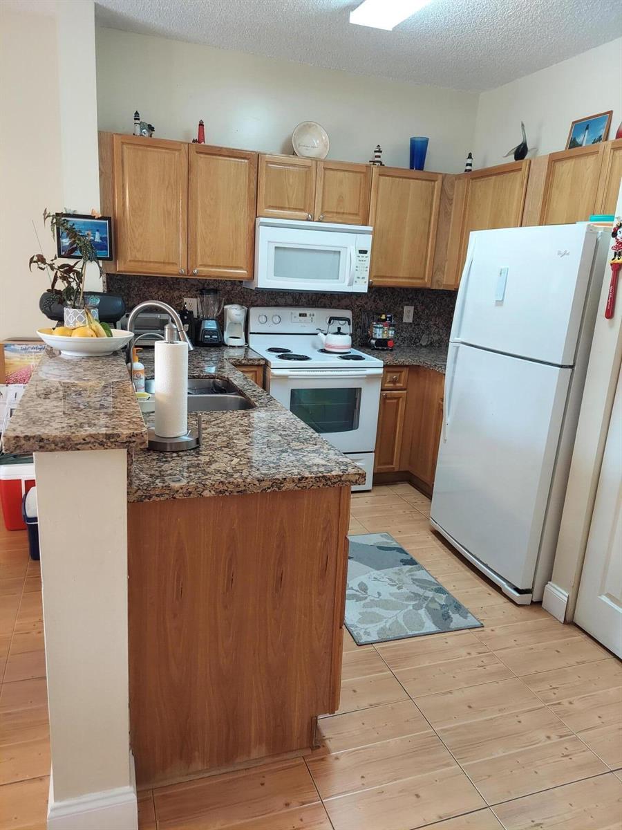 A kitchen features wood cabinets, granite countertops, white appliances, and light-colored tile flooring — 2208 SE 24th Place, Homestead FL 33035, HOMESTEAD, FL