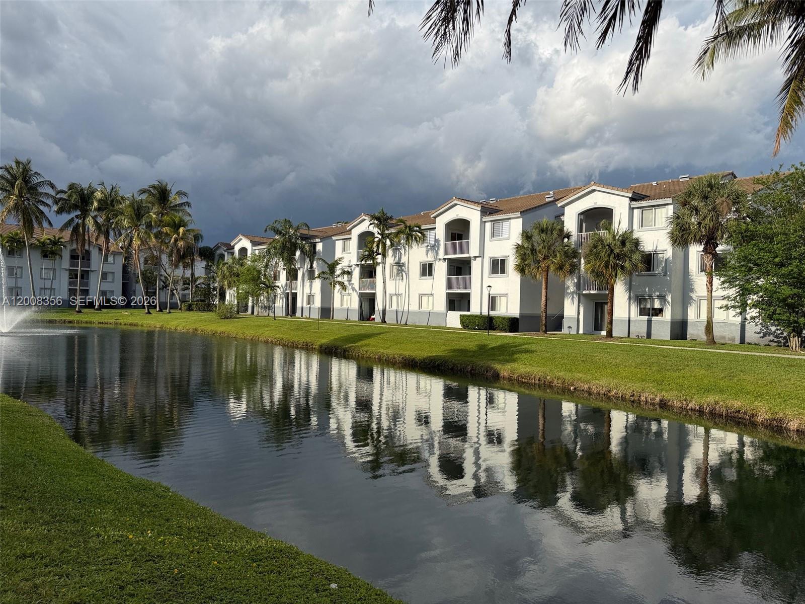 A multi-story apartment building sits beside a pond with palm trees and a fountain — 2600 S University Dr # 226, Davie FL 33328, DAVIE, FL