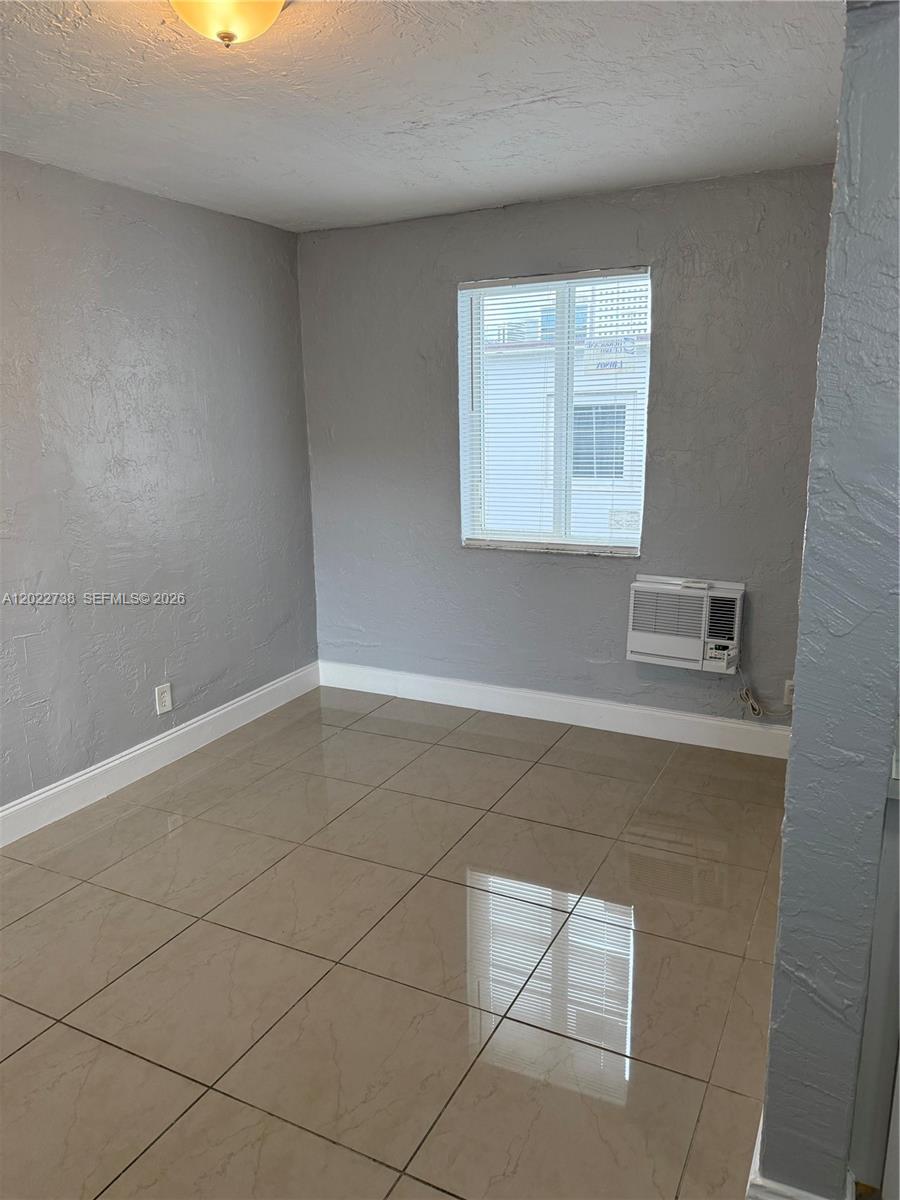 A room with gray textured walls, beige tile flooring, a window, and a wall-mounted air conditioner — MIAMI, FL
