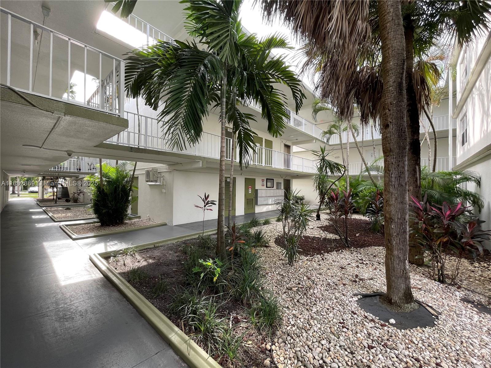 An outdoor courtyard features palm trees, gravel landscaping, and white multi-story apartment building walkways — 4255 N University Dr # 201, Sunrise FL 33351, SUNRISE, FL