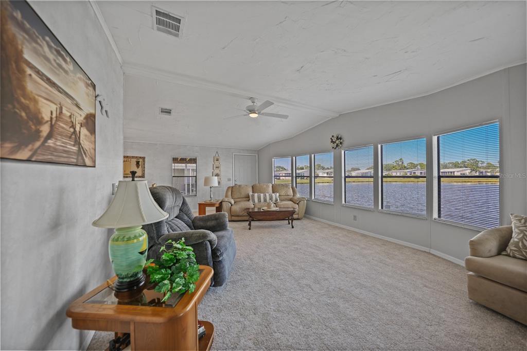 A carpeted living room features a ceiling fan, neutral walls, and large windows overlooking water — 495 SCHOONER STREET, NORTH PORT, FL