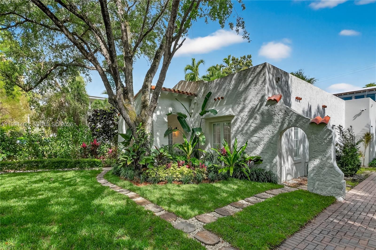 A white stucco house with a tiled roof and arched entryway sits behind a garden — 1109 Almeria Ave, Coral Gables FL 33134, CORAL GABLES, FL