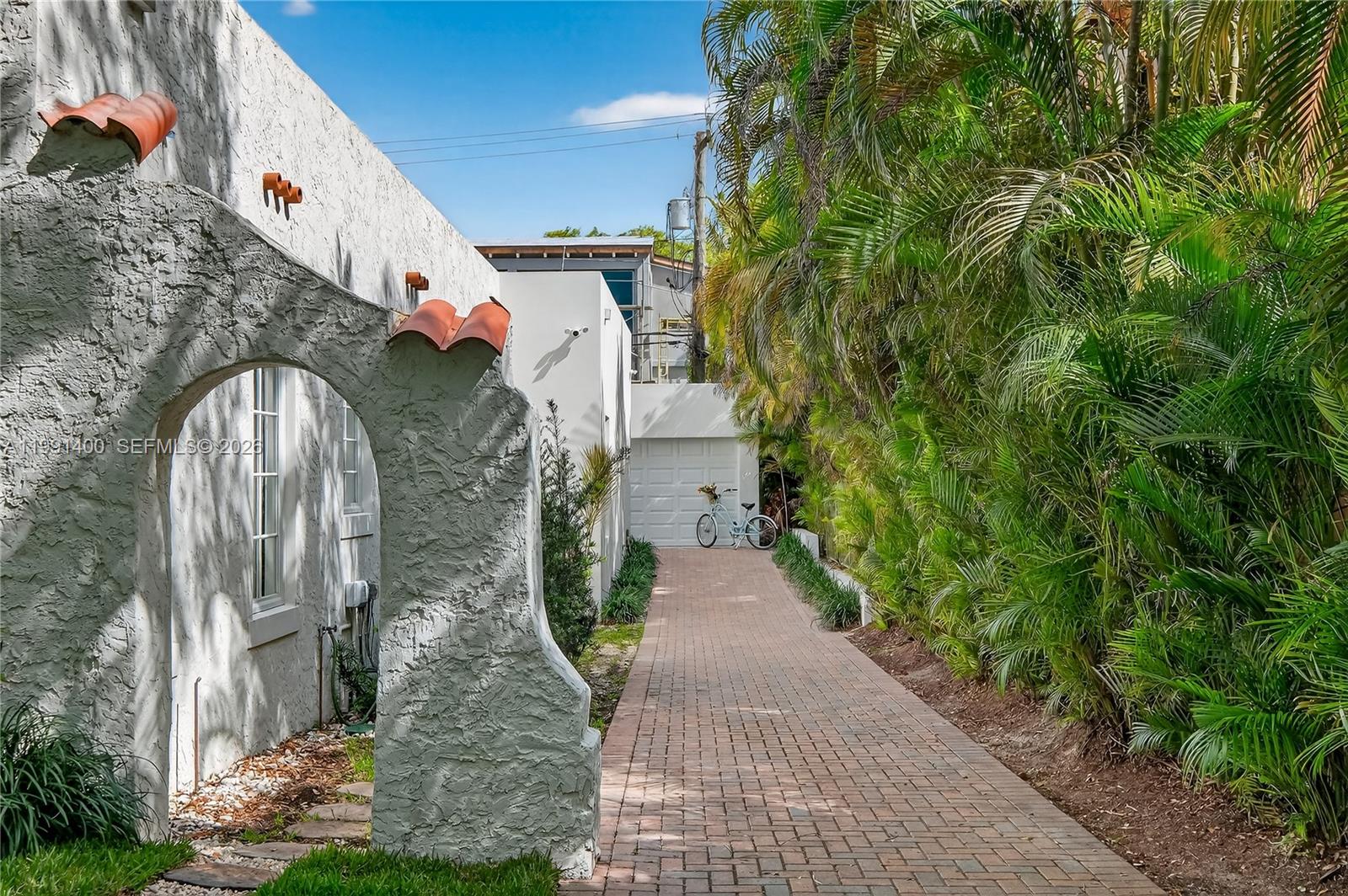 A paved walkway leads past a stucco archway toward a white garage and palm trees — 1109 Almeria Ave, Coral Gables FL 33134, CORAL GABLES, FL