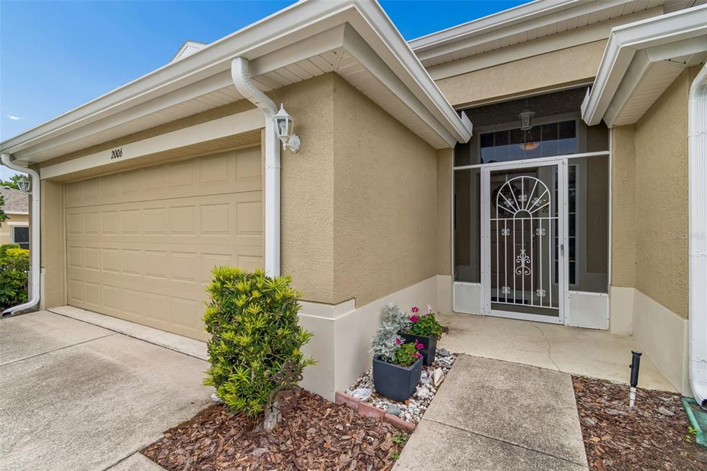 A beige house exterior features a two-car garage, a screened front door, and potted plants — 2006 GRANTHAM GREENS DRIVE UNIT 0, SUN CITY CENTER, FL