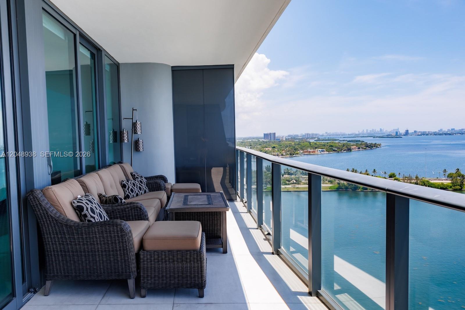 A balcony with wicker furniture overlooks a body of water and a distant city skyline — 3131 NE 7th Ave # 2003, Miami FL 33137, MIAMI, FL