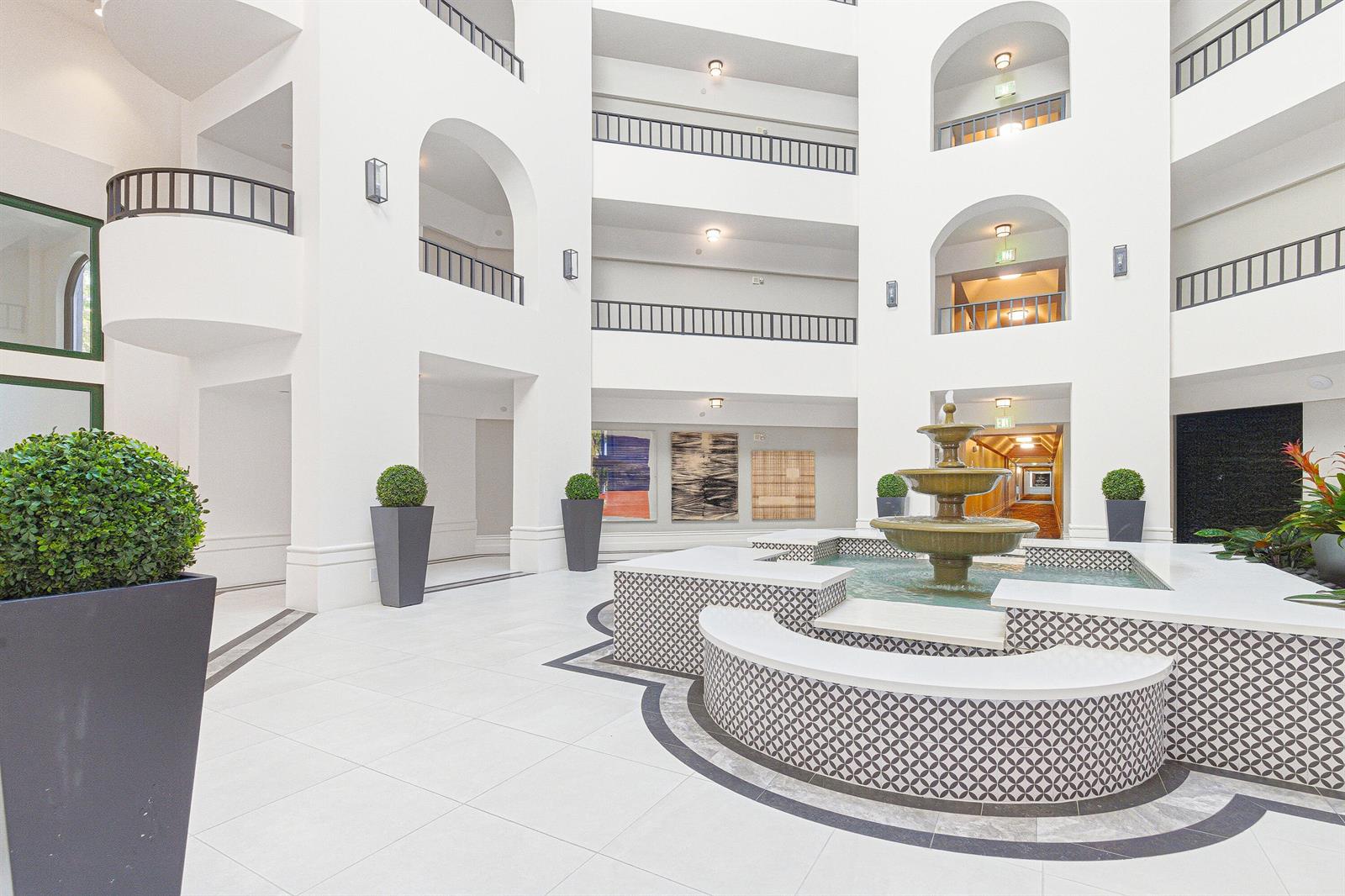 A multi-story atrium features a central tiered fountain, white tile flooring, and arched balconies — 100 SE 5th Avenue # 412, Boca Raton FL 33432, BOCA RATON, FL