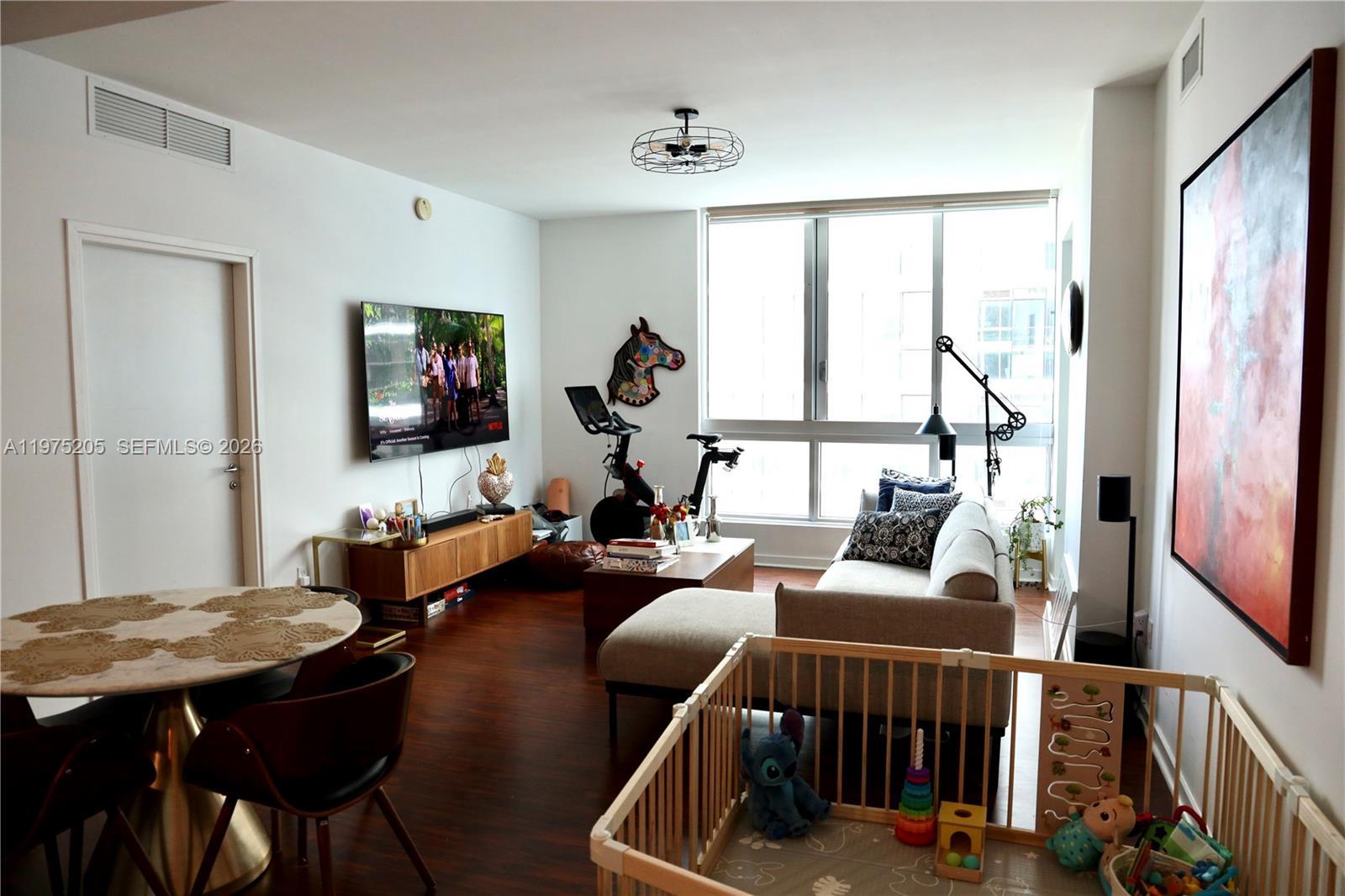 A living room features a sectional sofa, exercise bike, dining table, and a baby playpen — 300 S Biscayne Blvd # 3504, Miami FL 33131, MIAMI, FL