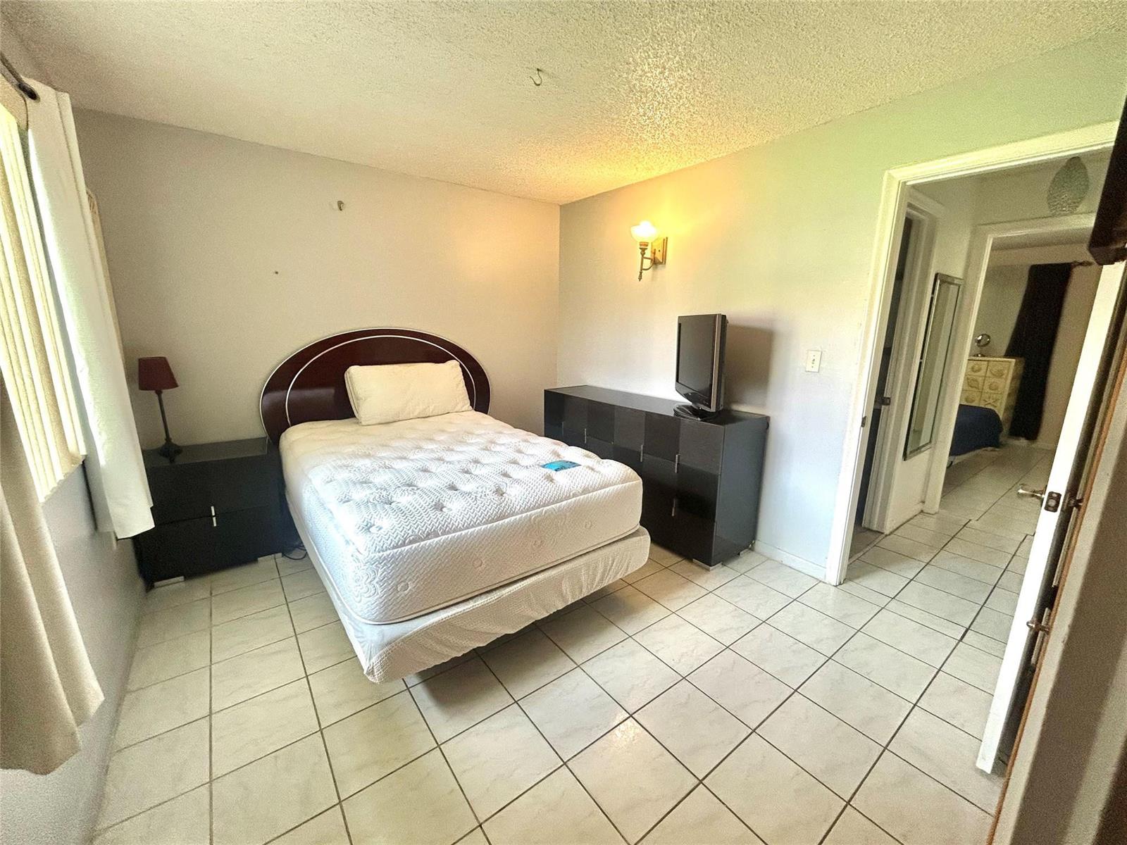 A bedroom features a bed, a dresser with a television, and white tile flooring — 6100 Forest Hill Boulevard # 101, West Palm Beach FL 33415, WEST PALM BEACH, FL