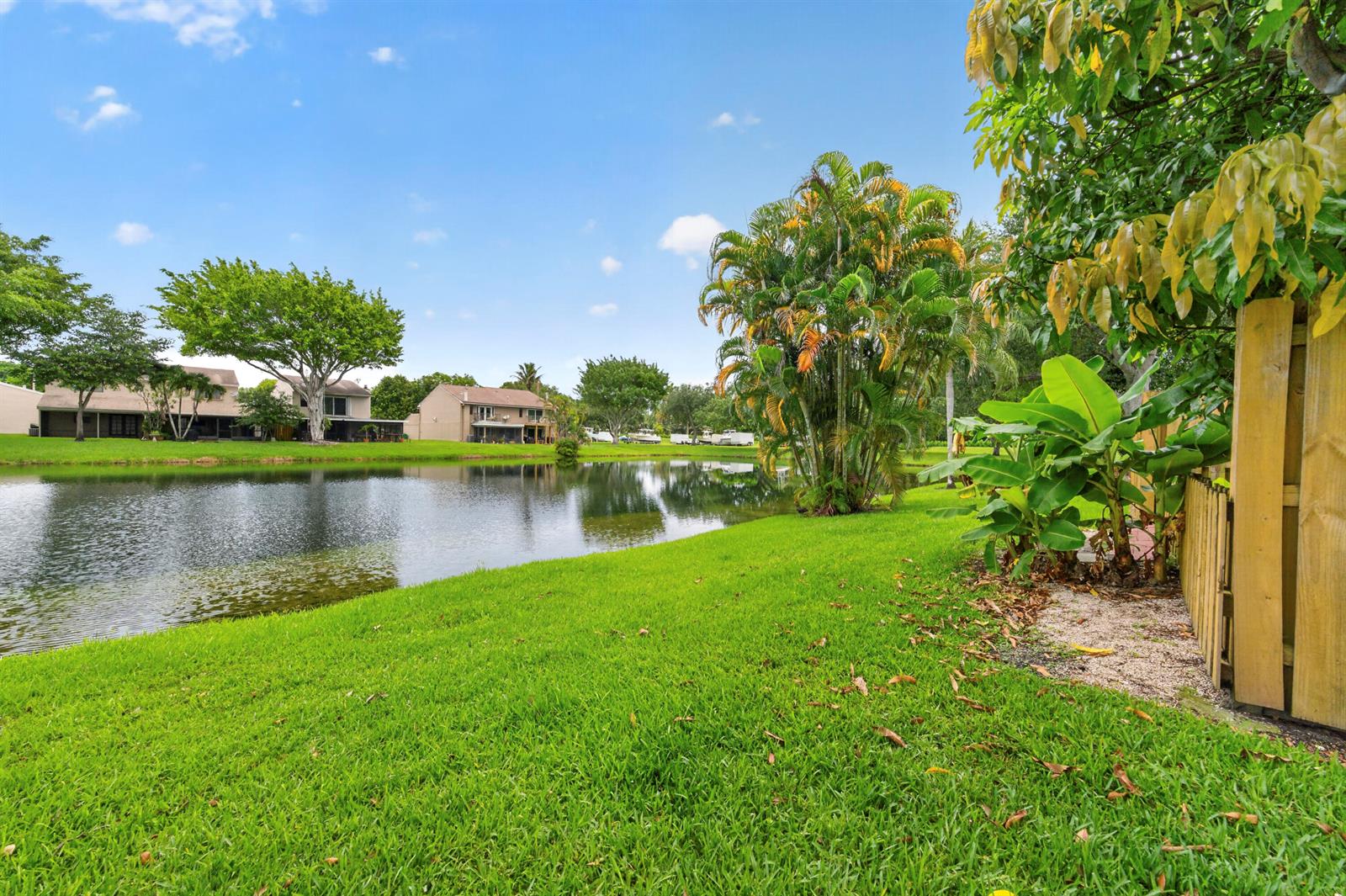 A grassy backyard overlooks a calm pond with palm trees and houses in the distance — 5182 S University Drive, Davie FL 33328, DAVIE, FL
