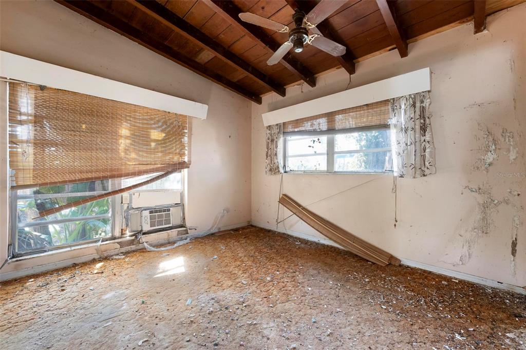 A bedroom with a wood-beamed ceiling, damaged walls, and exposed subflooring under two windows — 2956 GREENBRIAR STREET, SARASOTA, FL