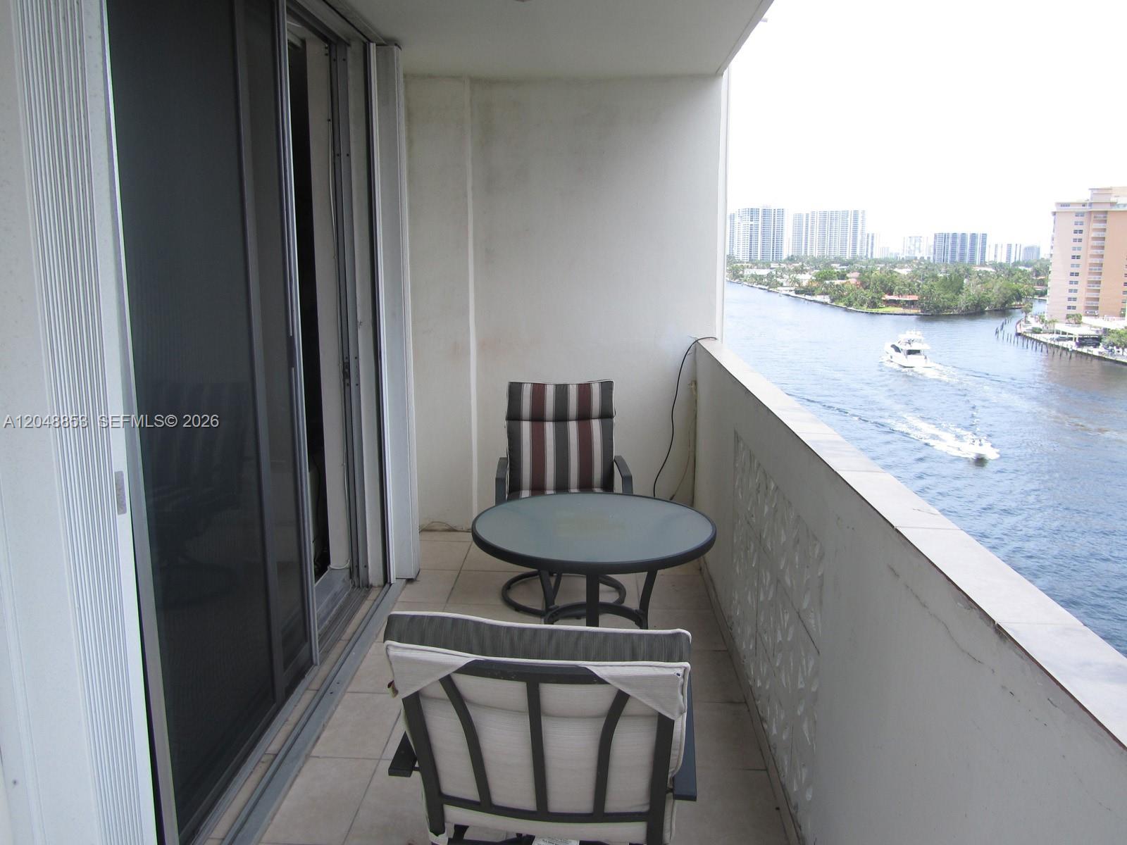 A balcony features a small glass table, two chairs, and a view of a waterway — 1833 S Ocean Dr # 911, Hallandale Beach FL 33009, HALLANDALE BEACH, FL