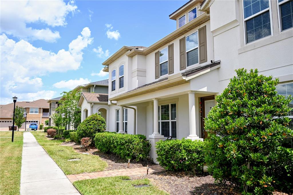 A multi-story townhouse with a covered front porch, tan stucco siding, and landscaped bushes — 2479 TOSCOLONA DRIVE, KISSIMMEE, FL