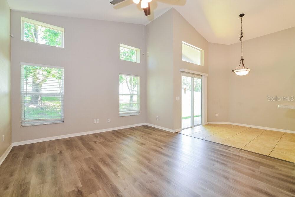 A living room with wood-look flooring, high ceilings, multiple windows, and a tiled dining area — 23 PATMORE ASH WAY, APOPKA, FL