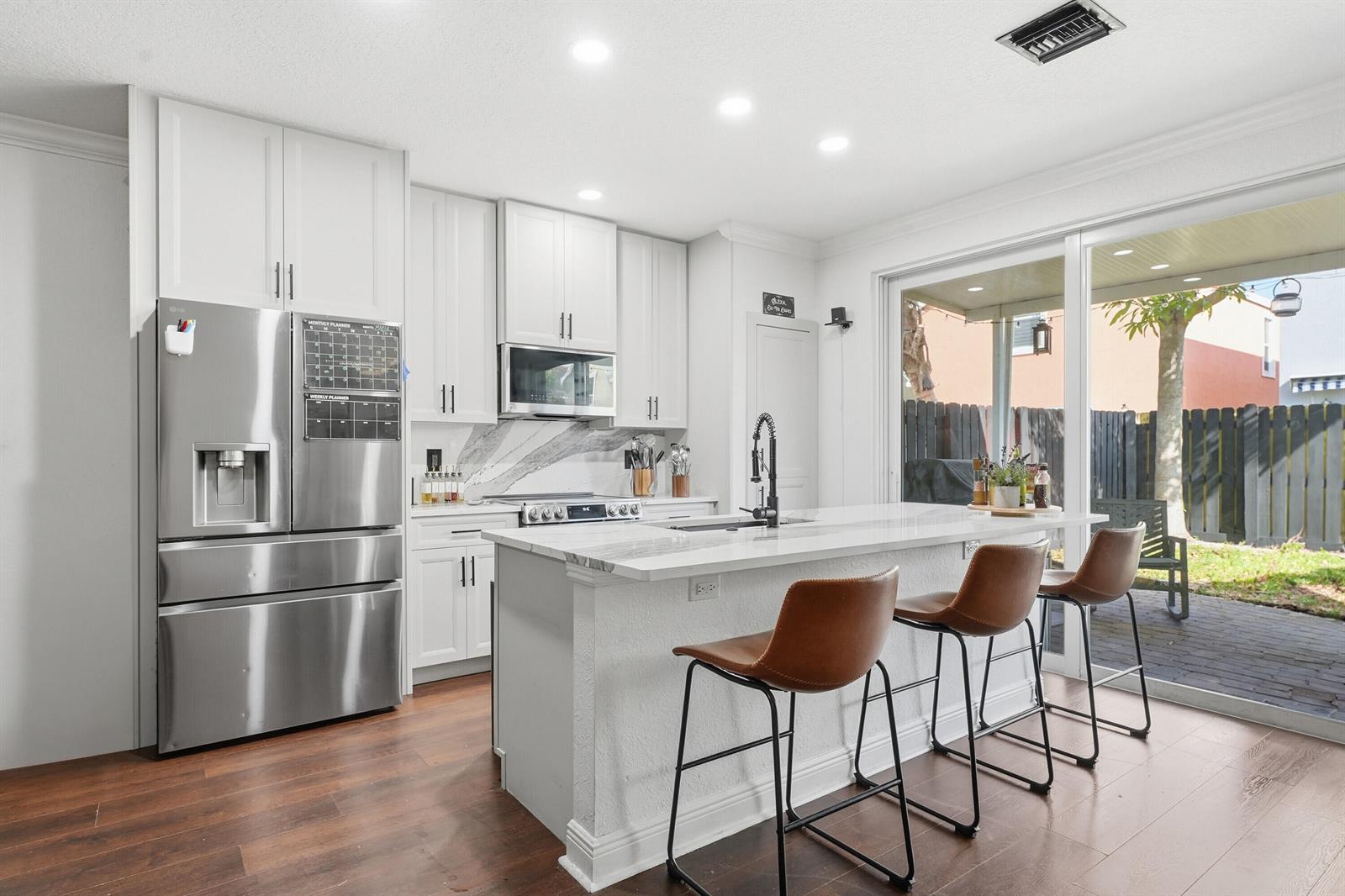 A modern kitchen features white cabinets, a marble-topped island with bar stools, and sliding doors — 3221 John's Place, Palm Springs FL 33461, PALM SPRINGS, FL