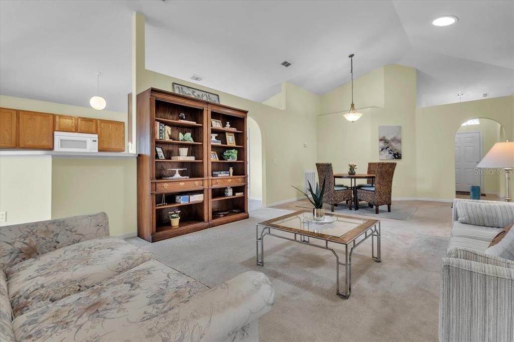 A living room and dining area with carpeted floors, a large wooden bookcase, and vaulted ceilings — 24483 LAKESIDE MANOR, PORT CHARLOTTE, FL