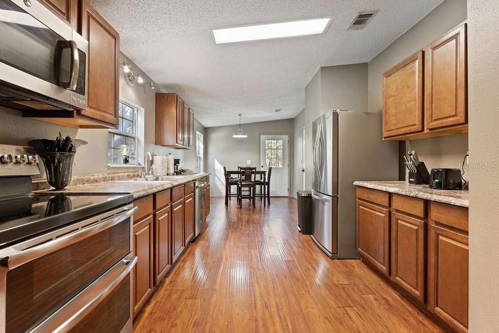 A galley kitchen with wood cabinets, stainless steel appliances, and wood-look flooring leads to dining — 8171 SE 144TH PLACE, INGLIS, FL