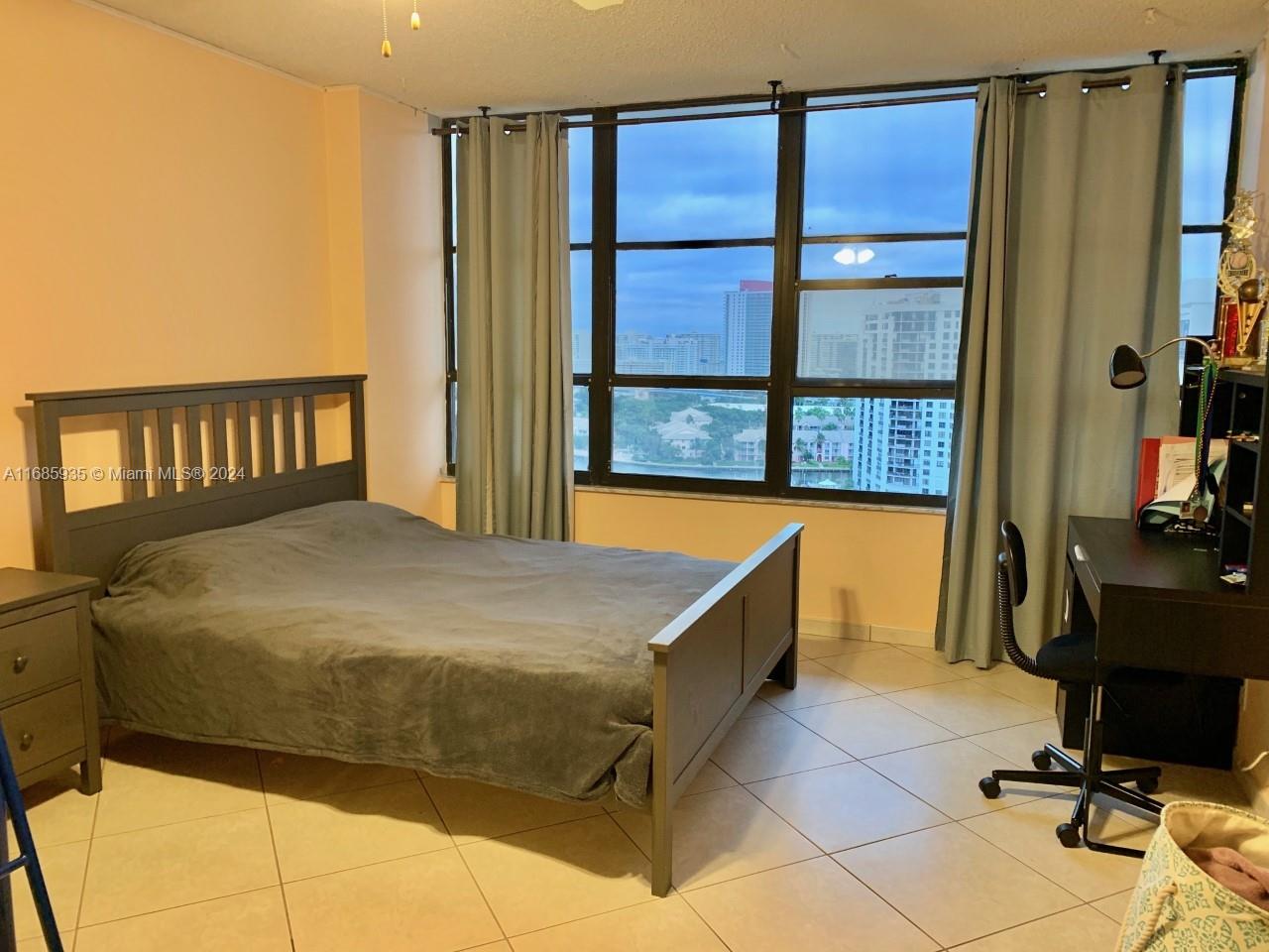 A bedroom features a gray bed frame, a desk, and large windows with city views — 600 Three Islands Blvd # 1817, Hallandale Beach FL 33009, HALLANDALE BEACH, FL