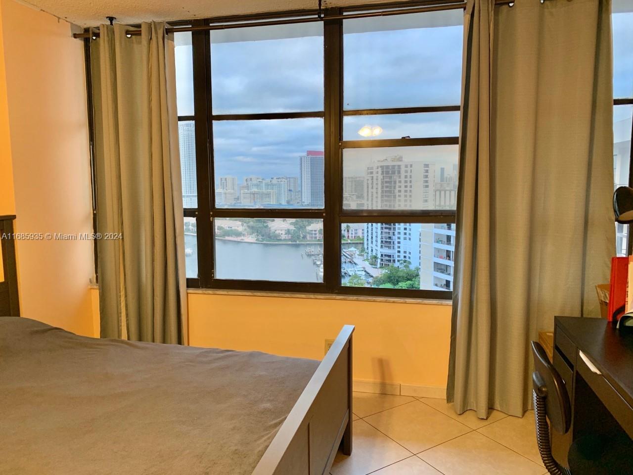 A bedroom features a bed, a desk, and large windows overlooking a waterfront city view — 600 Three Islands Blvd # 1817, Hallandale Beach FL 33009, HALLANDALE BEACH, FL
