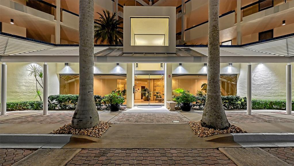 The exterior entrance of a multi-story building features a glass doorway, palm trees, and landscaping — 1932 HARBOURSIDE DRIVE UNIT 242, LONGBOAT KEY, FL