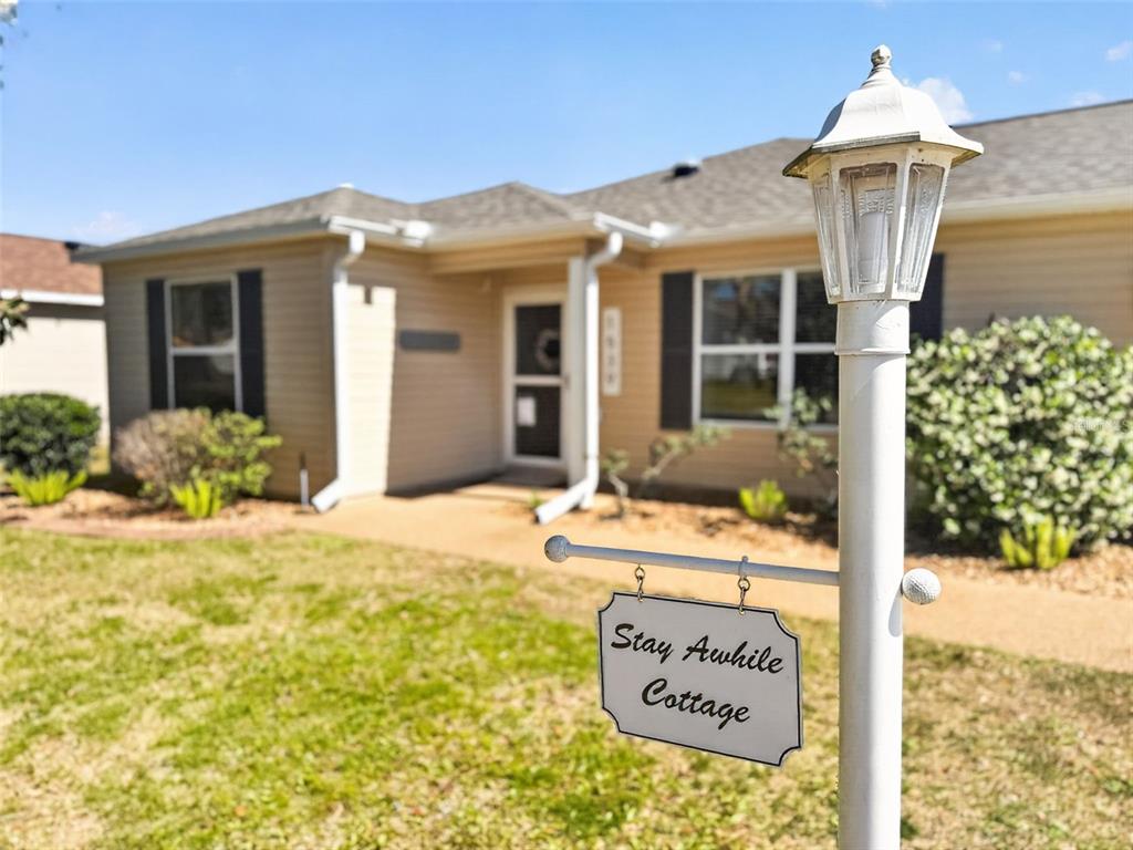 A beige house with a front lawn and a sign reading Stay Awhile Cottage — 1539 HOLLYBERRY PLACE, THE VILLAGES, FL