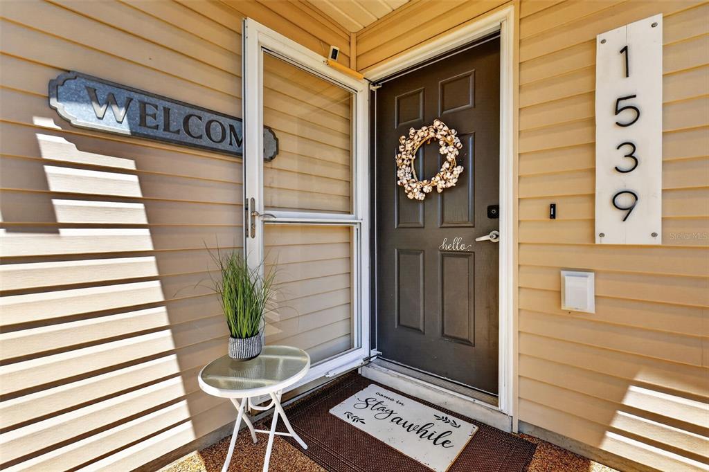 A front porch features a dark door with a wreath, a small table, and house numbers — 1539 HOLLYBERRY PLACE, THE VILLAGES, FL
