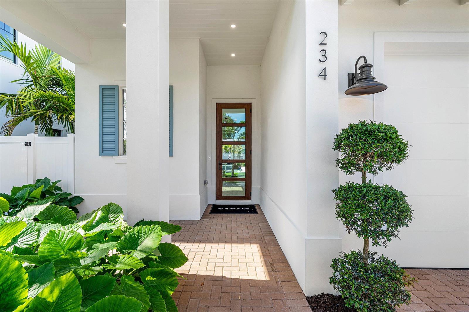A white exterior entryway features a wooden front door, brick pavers, and a topiary tree — 234 NE 12th Street, Delray Beach FL 33444, DELRAY BEACH, FL