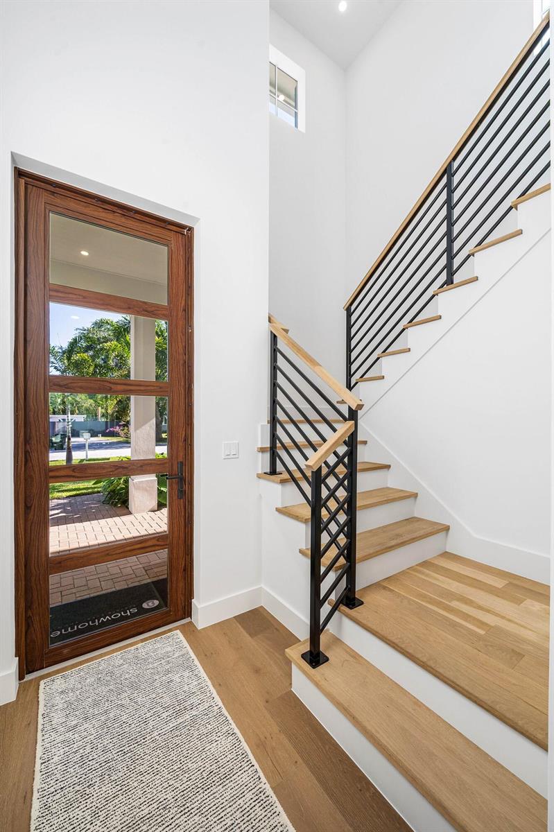 A modern entryway features a wooden front door, light wood flooring, and a black-railed staircase — 234 NE 12th Street, Delray Beach FL 33444, DELRAY BEACH, FL
