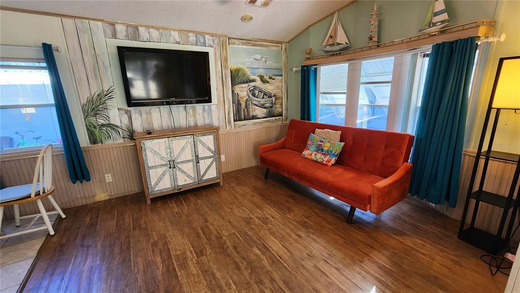 A living room features wood-look flooring, an orange sofa, a television, and coastal-themed wall art — 3300 26TH AVENUE E UNIT 225, BRADENTON, FL