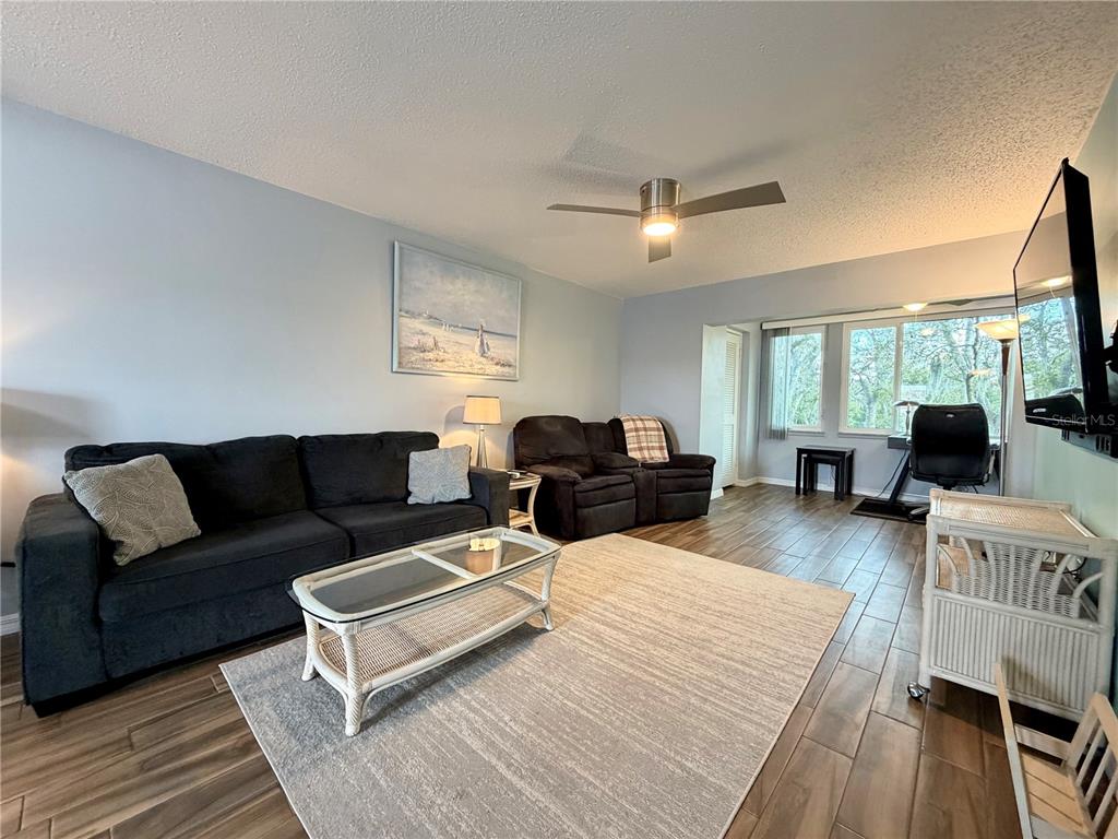 A living room features a dark sofa, a recliner, a glass coffee table, and wood-look flooring — 4807 MARINE PARKWAY UNIT 202, NEW PORT RICHEY, FL