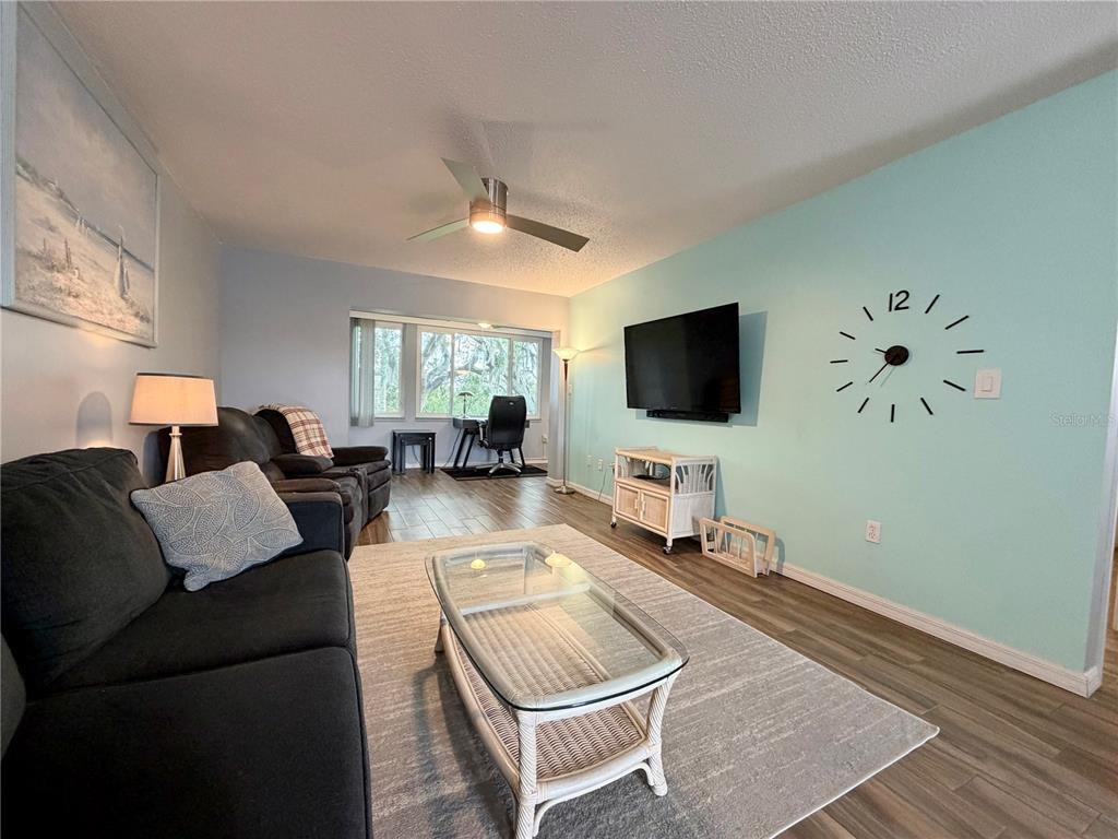 A living room features a dark sofa, glass coffee table, wall-mounted clock, and a workspace area — 4807 MARINE PARKWAY UNIT 202, NEW PORT RICHEY, FL