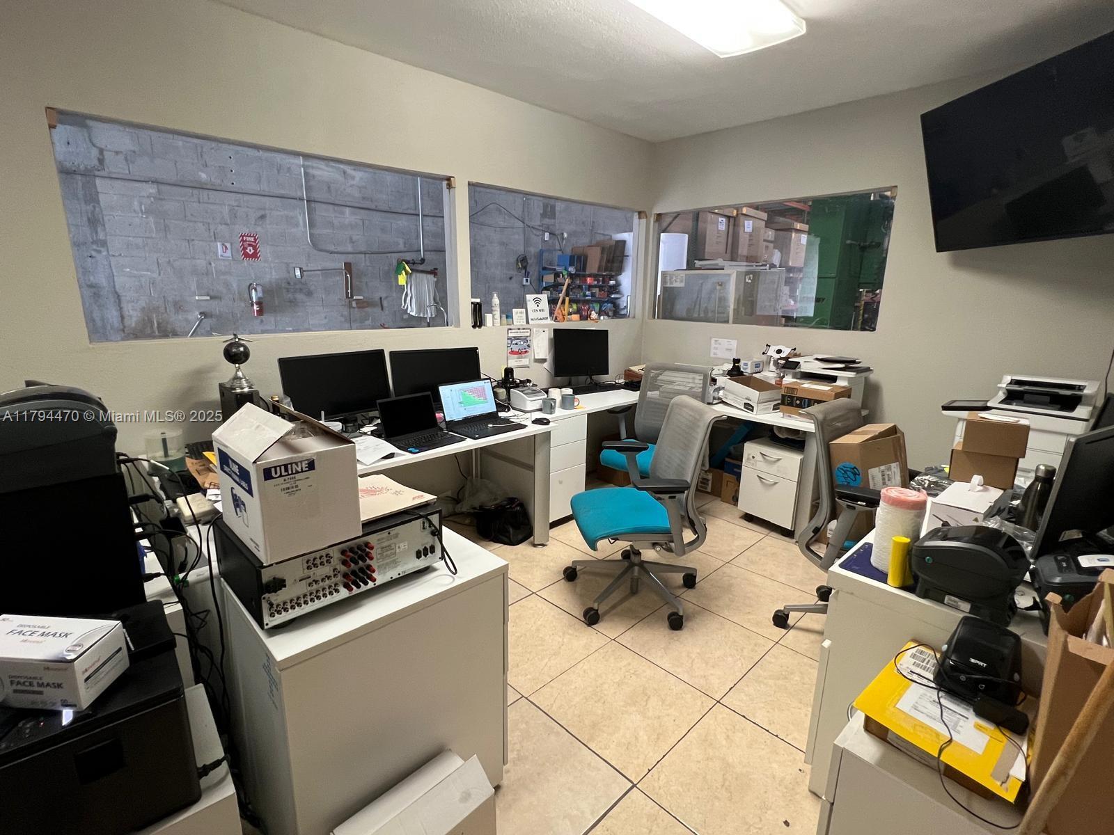 An office space features multiple desks, computer equipment, office chairs, and windows overlooking a warehouse — 2330 NW 102nd Pl, Doral FL 33172, DORAL, FL