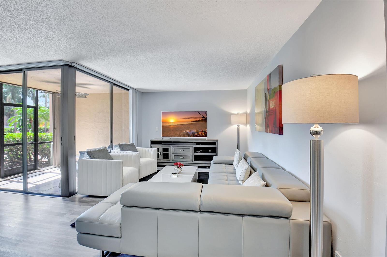 A modern living room features a grey sectional, white armchairs, a television, and sliding glass doors — 2255 Lindell Boulevard # 4103, Delray Beach FL 33444, DELRAY BEACH, FL