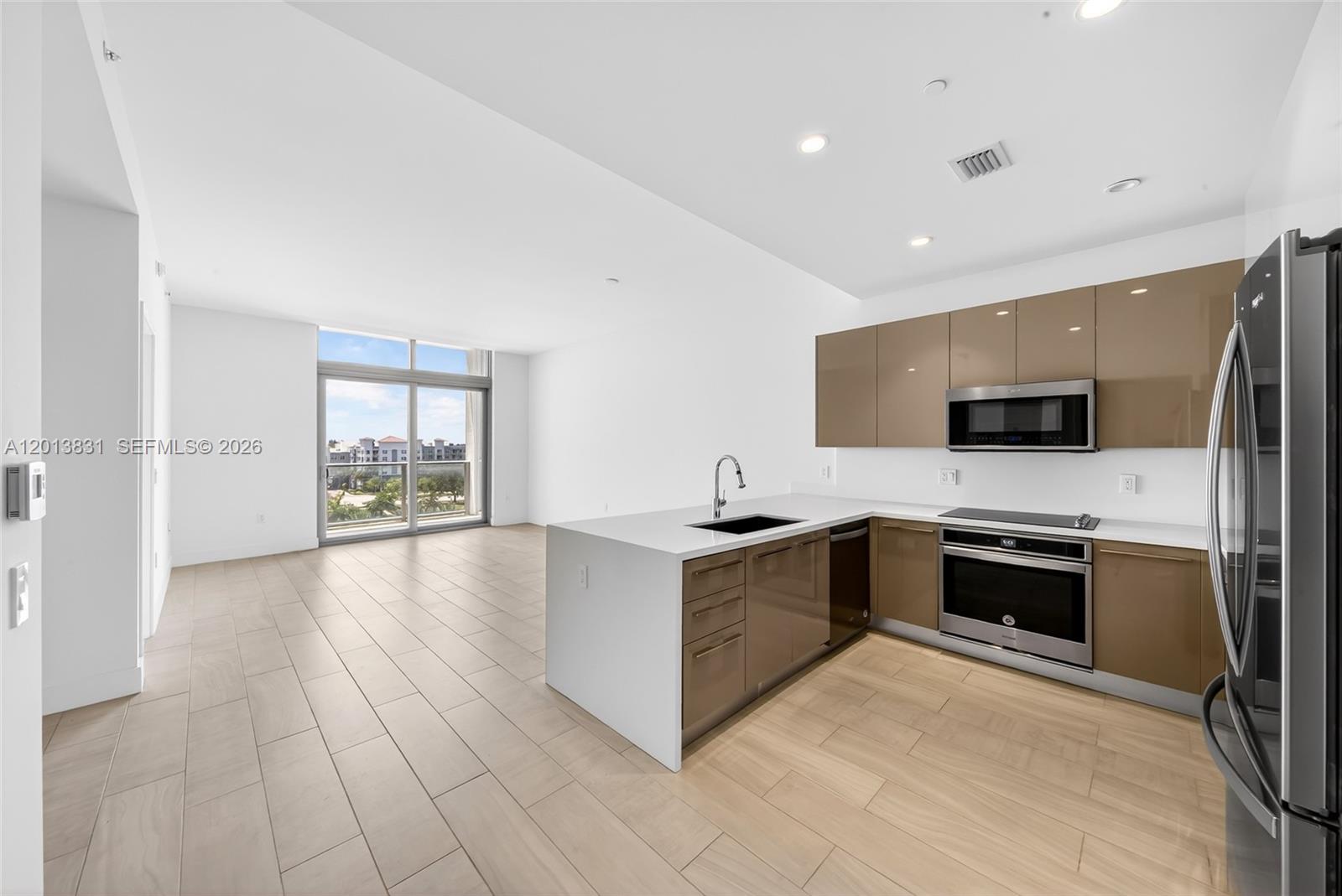 A modern kitchen with glossy brown cabinets and white countertops opens to a living area — 2000 Metropica Way # 615, Sunrise FL 33323, SUNRISE, FL
