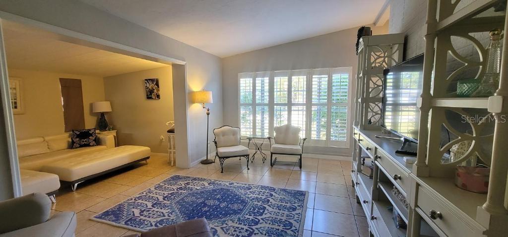 A living room features tile floors, a blue area rug, plantation shutters, and an entertainment center — 3530 SCHWALBE DRIVE, SARASOTA, FL