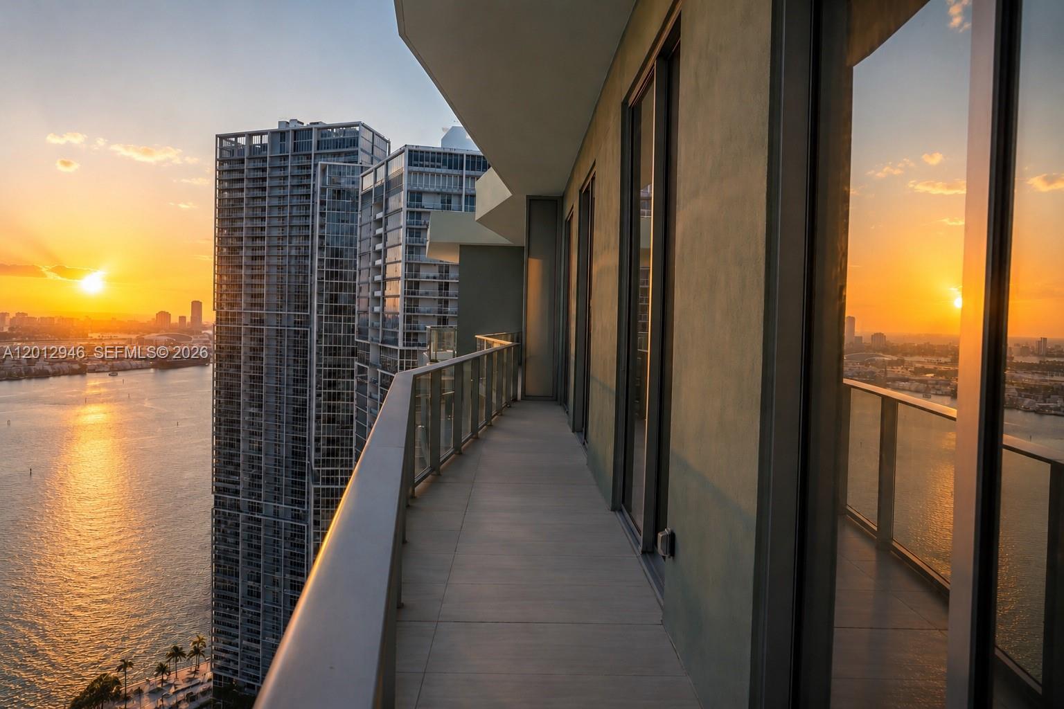 A high-rise balcony with glass railings overlooks a bay at sunset next to tall buildings — 77 SE 5 ST, Miami FL 33131, MIAMI, FL