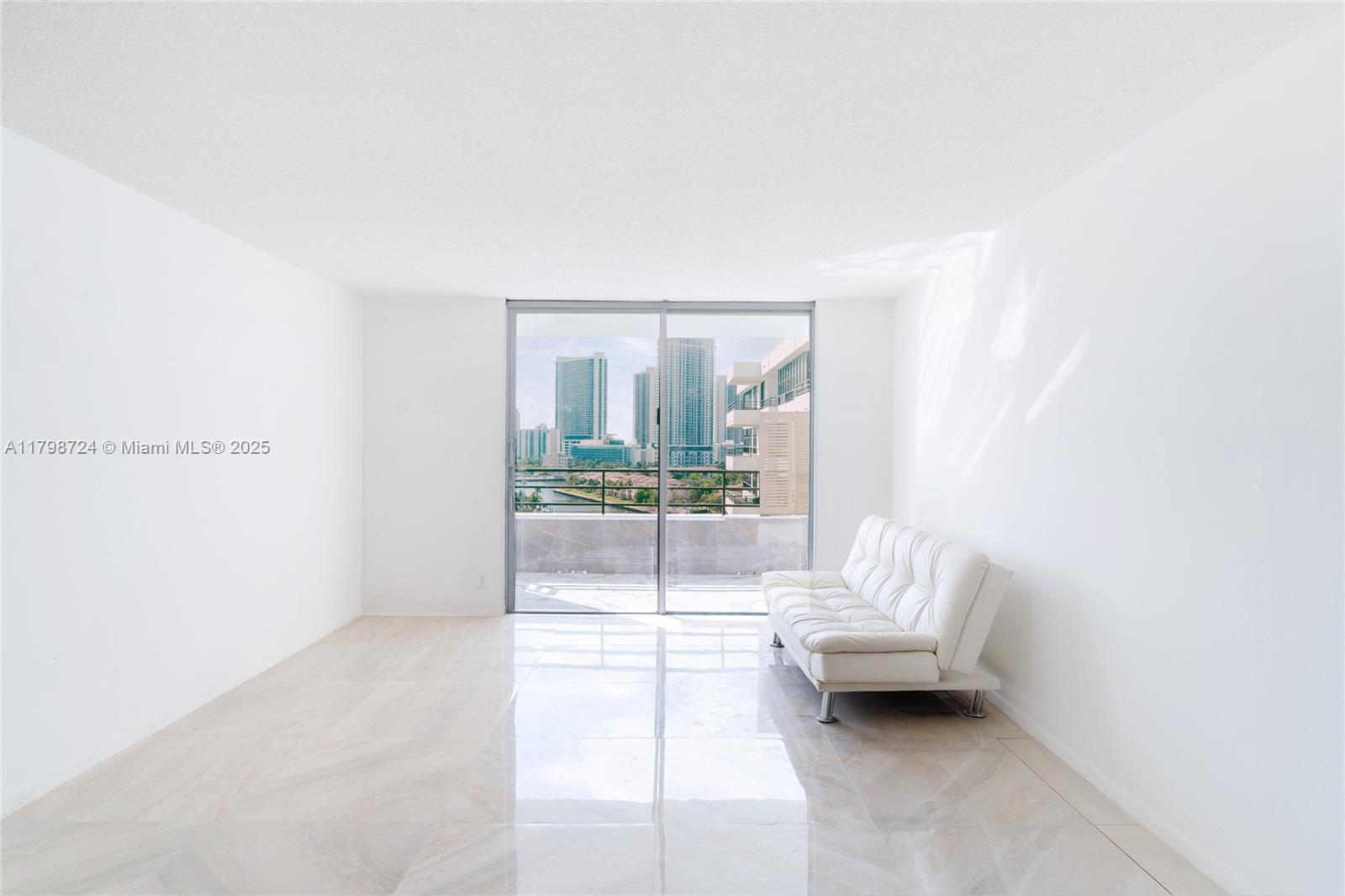A minimalist living room features white walls, light tile flooring, and a sliding glass balcony door — 500 Three Islands Blvd # 801, Hallandale Beach FL 33009, HALLANDALE BEACH, FL