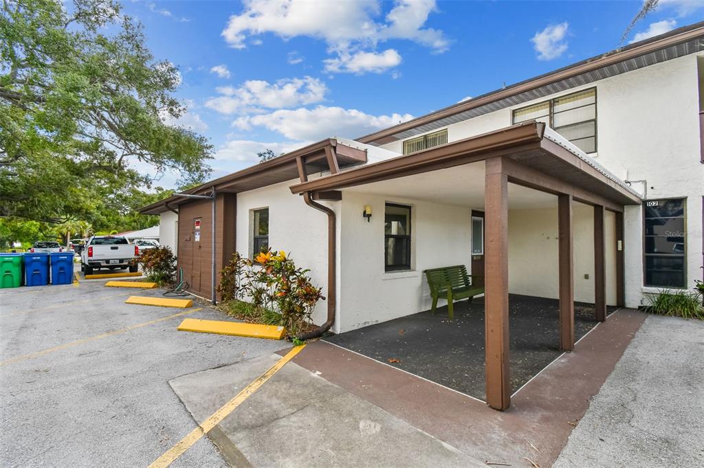 A white building exterior features a covered patio with a green bench and parking spaces — 8423 SEMINOLE BLVD UNIT 203, SEMINOLE, FL