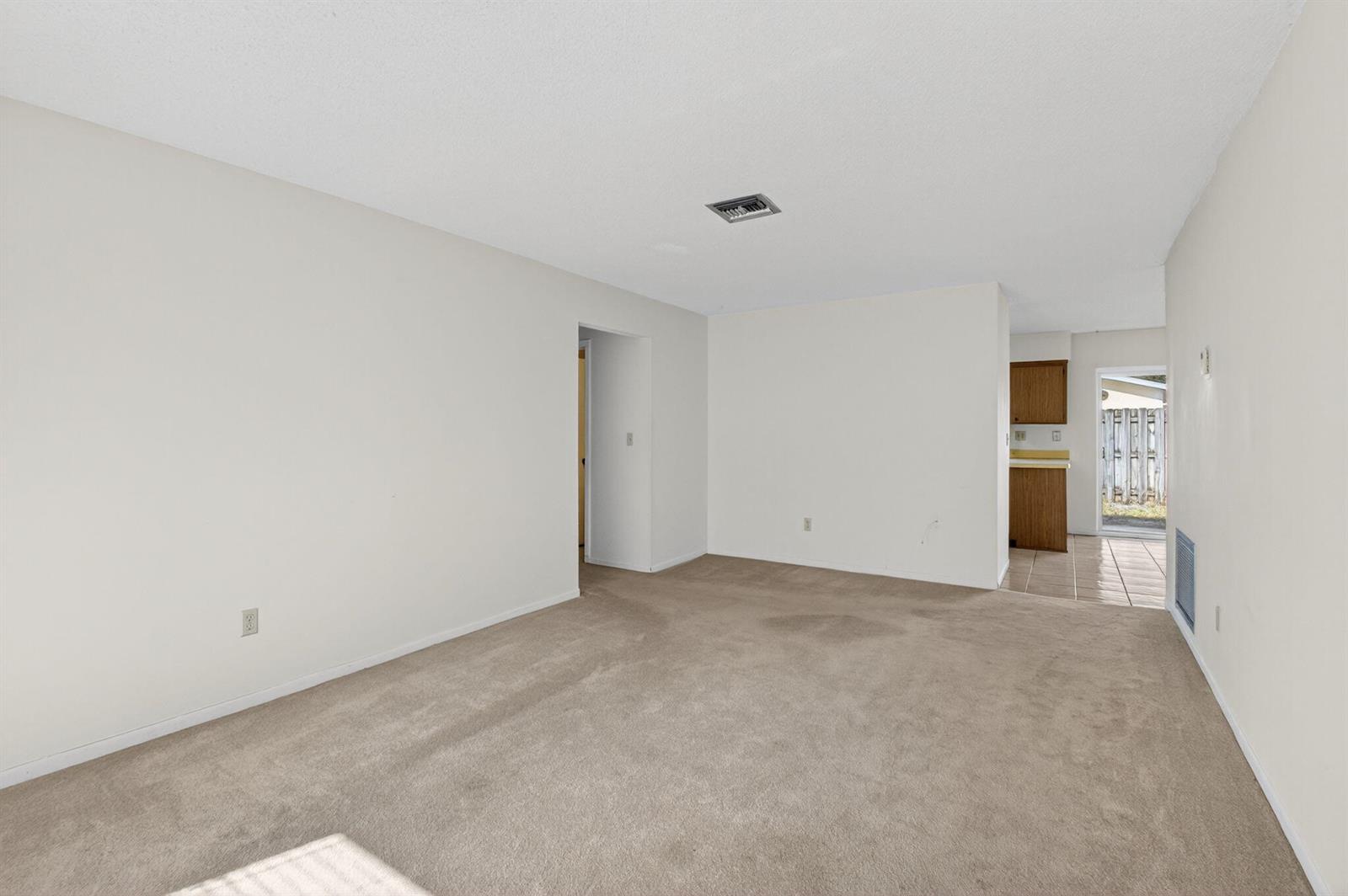 An empty living room with beige carpet, white walls, and a view into the kitchen — 1601 NW 5th Street, Boca Raton FL 33486, BOCA RATON, FL