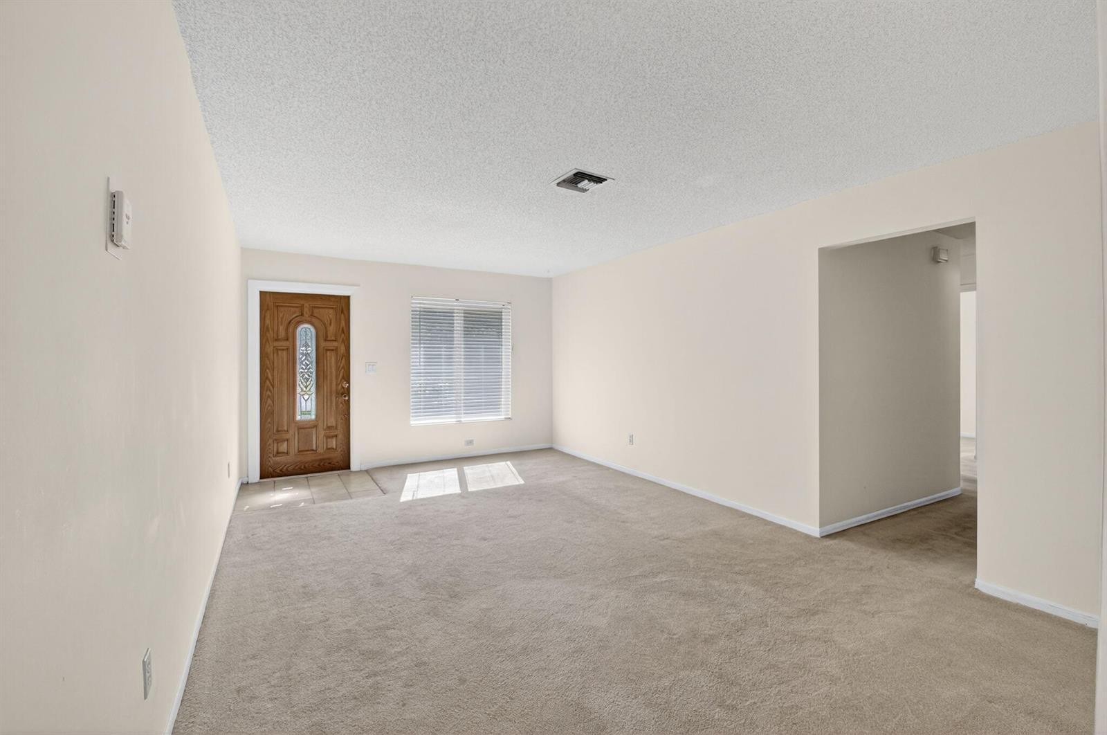 An empty living room features beige carpeting, light-colored walls, a front door, and a window — 1601 NW 5th Street, Boca Raton FL 33486, BOCA RATON, FL