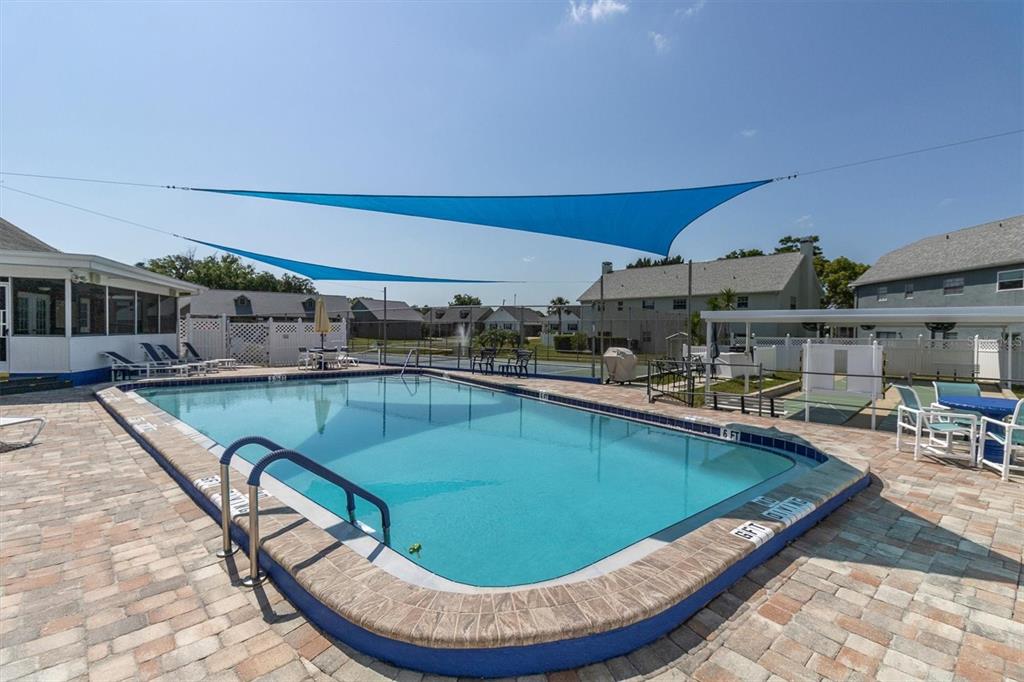 A rectangular swimming pool with a blue shade sail sits on a paved patio — 6129 ELMHURST DRIVE, NEW PORT RICHEY, FL