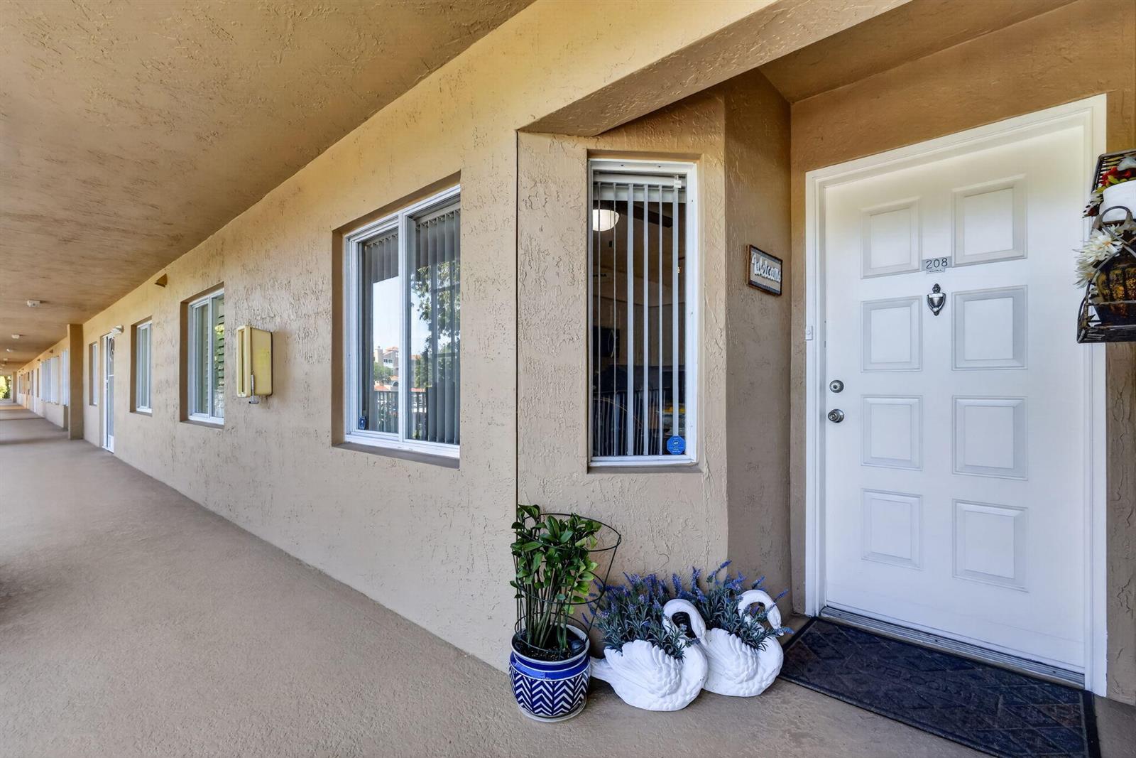 An exterior view of an apartment building walkway with a white door and potted plants — 5906 Crystal Shores Drive # 208, Boynton Beach FL 33437, BOYNTON BEACH, FL