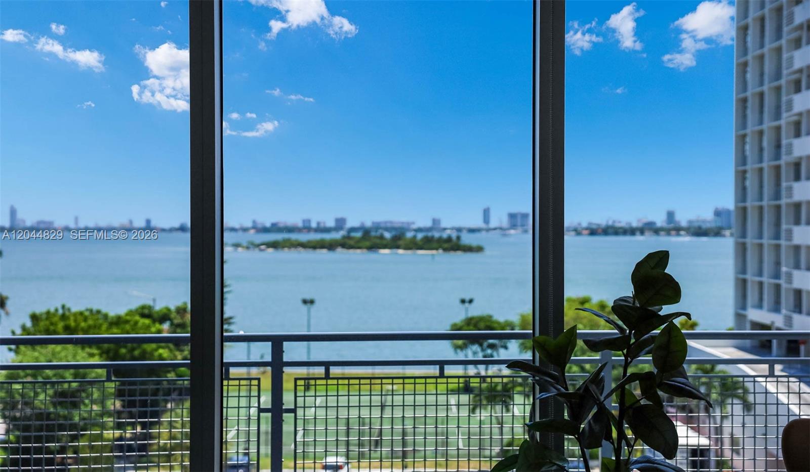 A view of a bay and city skyline through a window with a balcony railing — 2150 N Bayshore Dr # 1602, Miami FL 33137, MIAMI, FL