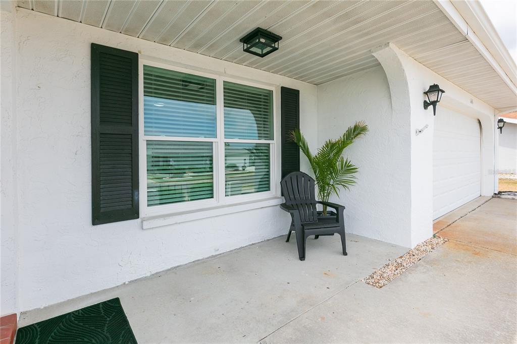 A covered concrete porch features a black chair, a potted plant, and a window — 1616 BENTWOOD DRIVE, SUN CITY CENTER, FL