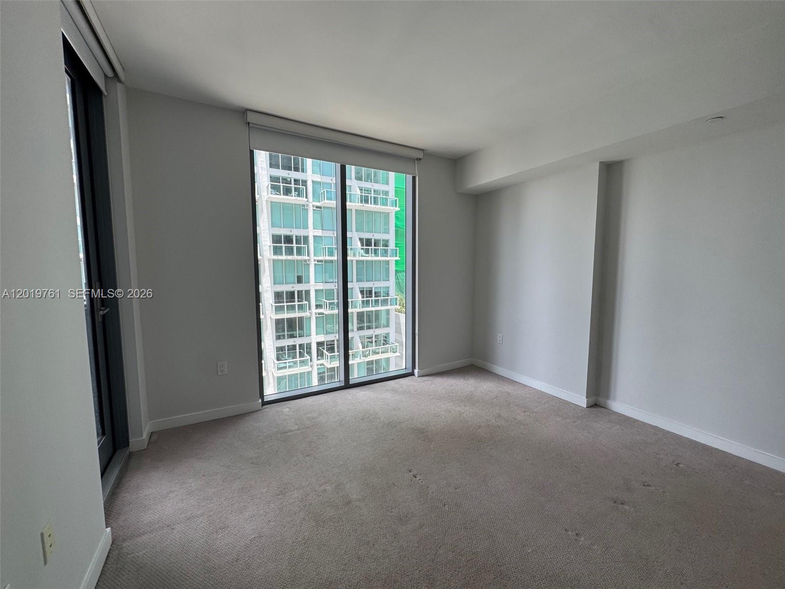 An empty room features beige carpeting, white walls, and a large window overlooking another building — 31 SE 6th St # 2004, Miami FL 33131, MIAMI, FL