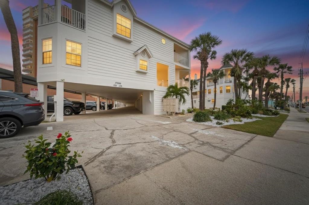 A white multi-story building with elevated parking sits beside a palm-lined street at sunset — 17960 GULF BOULEVARD UNIT 119, REDINGTON SHORES, FL